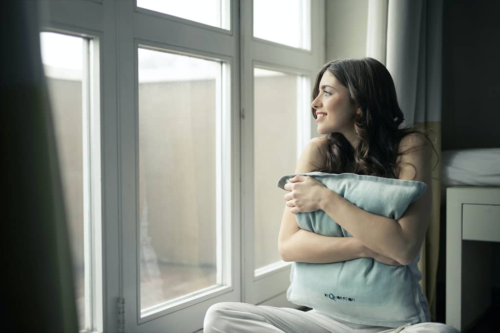 Dark haired woman hugging a pillowcase and smiling while looking out the window during her mental health day