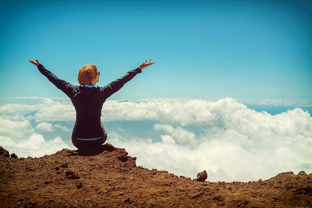 Person sitting on a cliff with both arms raised in gratitude