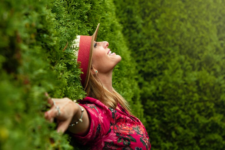 A woman with a hat leaning against tall bushes smiling