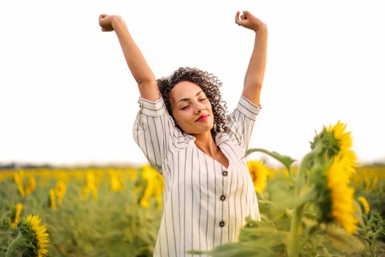 A woman raising her arms in the air with her eyes closed in a field of sunflowers