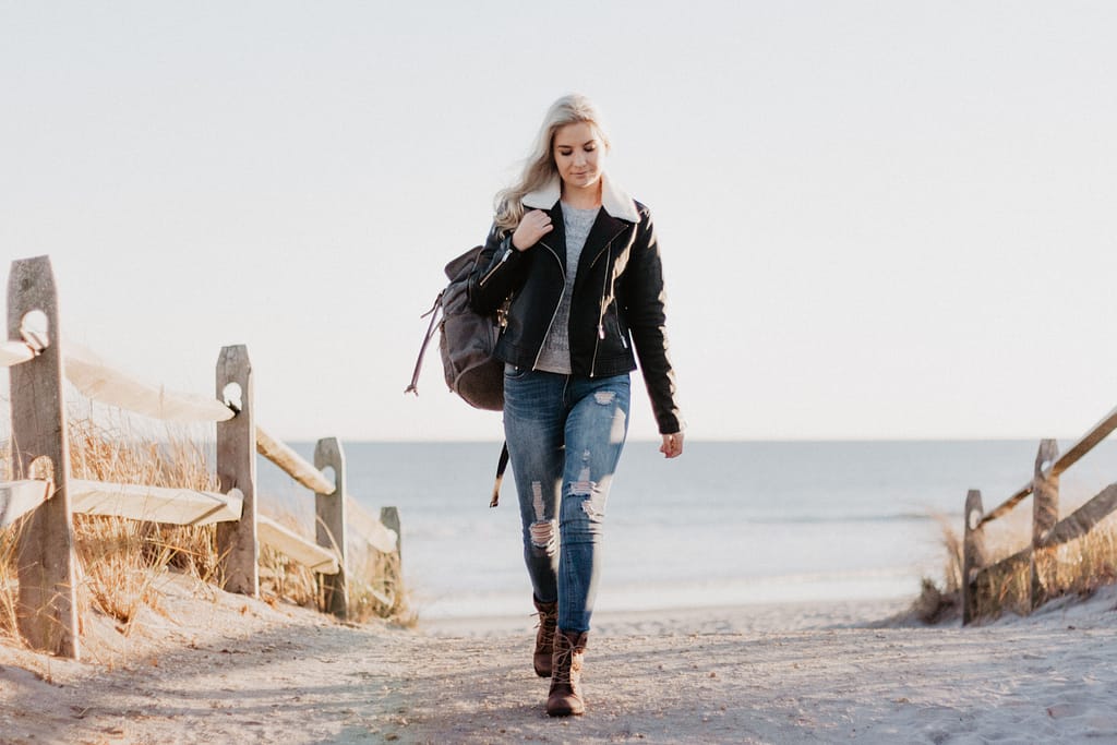 A young woman with long hair wearing jeans, boots and a leather jacket with a backpack walking near the beach