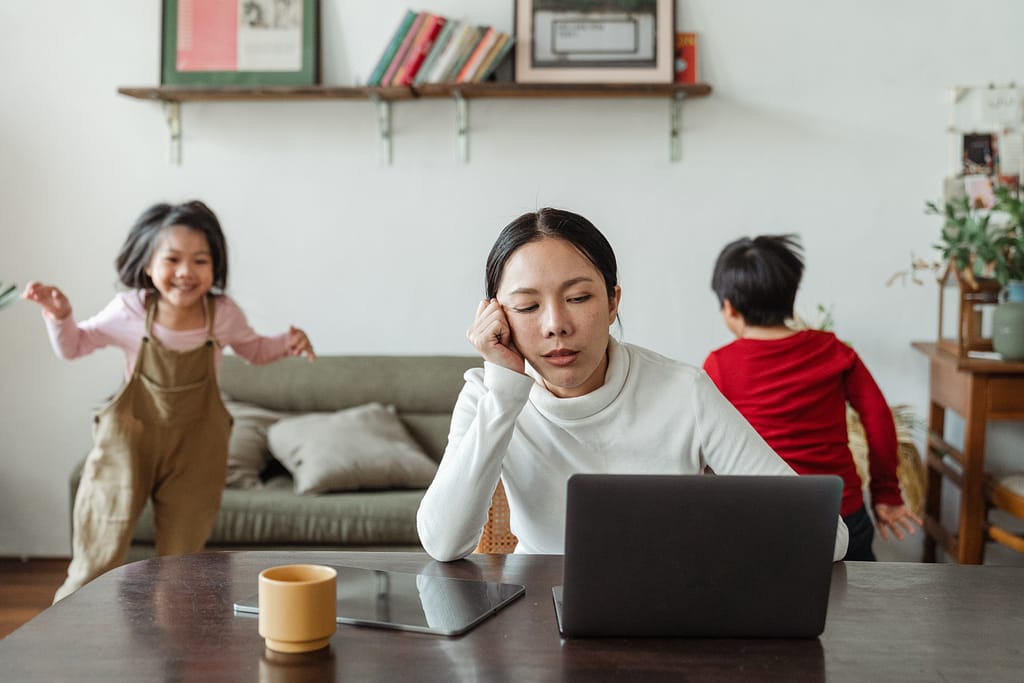A woman looking at a computer while her children play in the background