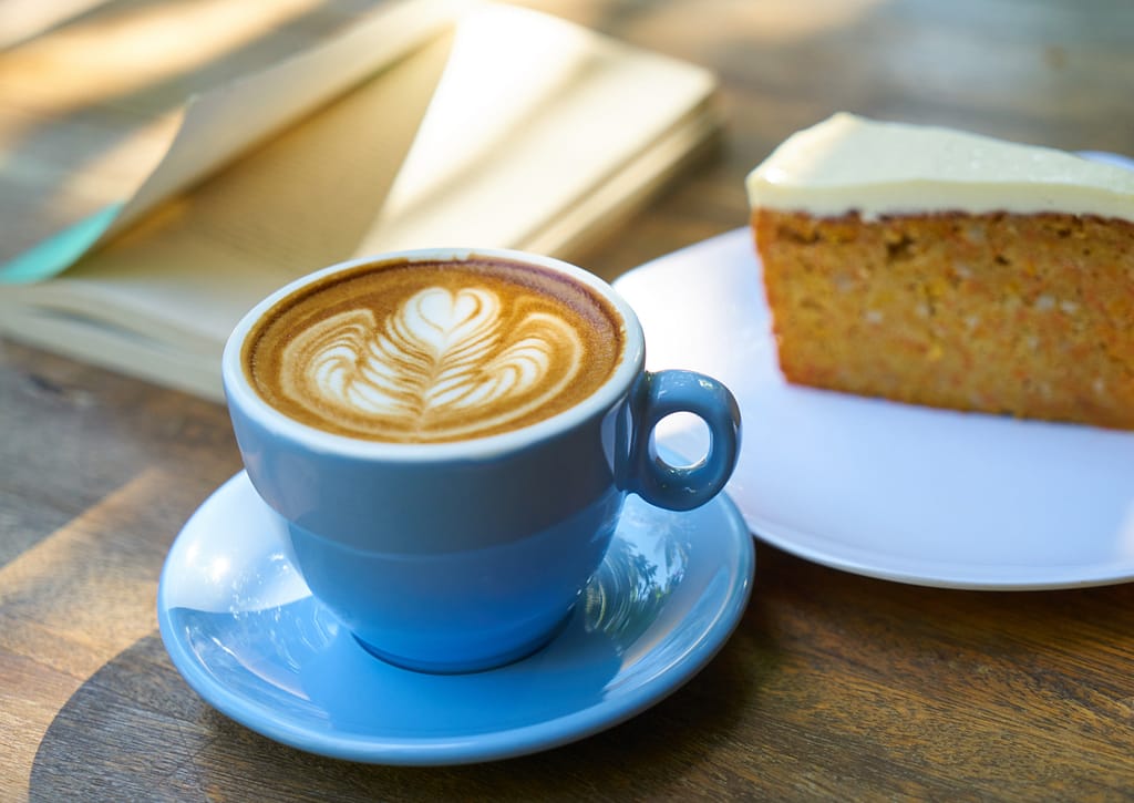 A latte with an intricate design on top next to a slice of carrot cake