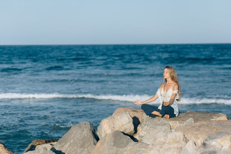 A woman meditating while sitting on rocks by the ocean