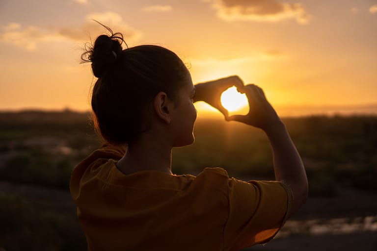 A woman facing the sunset with her fingers making a shape of a heart